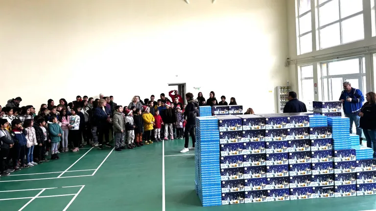 A group of children and adults gather in a brightly lit gymnasium for a gift distribution event. The children stand in a line near a large stack of festive blue and white boxes featuring holiday-themed designs. Several adults oversee the event, with one person addressing the group. Large windows provide natural light, enhancing the open and welcoming atmosphere.