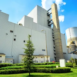 An industrial power plan with a modern design, featuring a large white building with corrugated metal siding and a tall concrete smokestack. A cooling tower is visible in the background. The foreground includes a well-maintained green lawn, small bushes, and a young pine tree under a bright blue sky with scattered clouds.