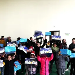 A group of smiling children in a gymnasium holding up blue and white holiday-themed gift boxes with festive designs. Many children are dressed in warm winter clothing, some wearing Santa hats. The joyful atmosphere is evident as they pose with their gifts. Adults in the background are observing the event, creating a lively and celebratory scene in a well-lit indoor space.